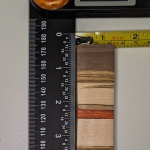 May include: A wooden chopping board with a multi-coloured wood grain pattern. The board is approximately 1.5 inches wide and 4 inches long. The board is measured with a ruler and a tape measure.