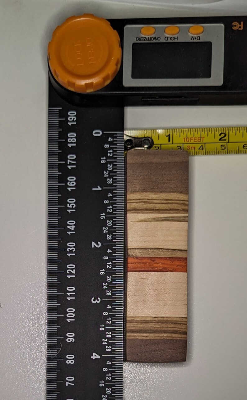 May include: A wooden chopping board with a multi-coloured wood grain pattern. The board is approximately 1.5 inches wide and 4 inches long. The board is measured with a ruler and a tape measure.
