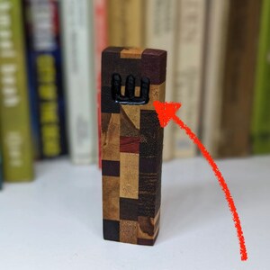 May include: A wooden mezuzah with a black Hebrew letter Shin on the front. The mezuzah is made of different types of wood, creating a patchwork pattern.