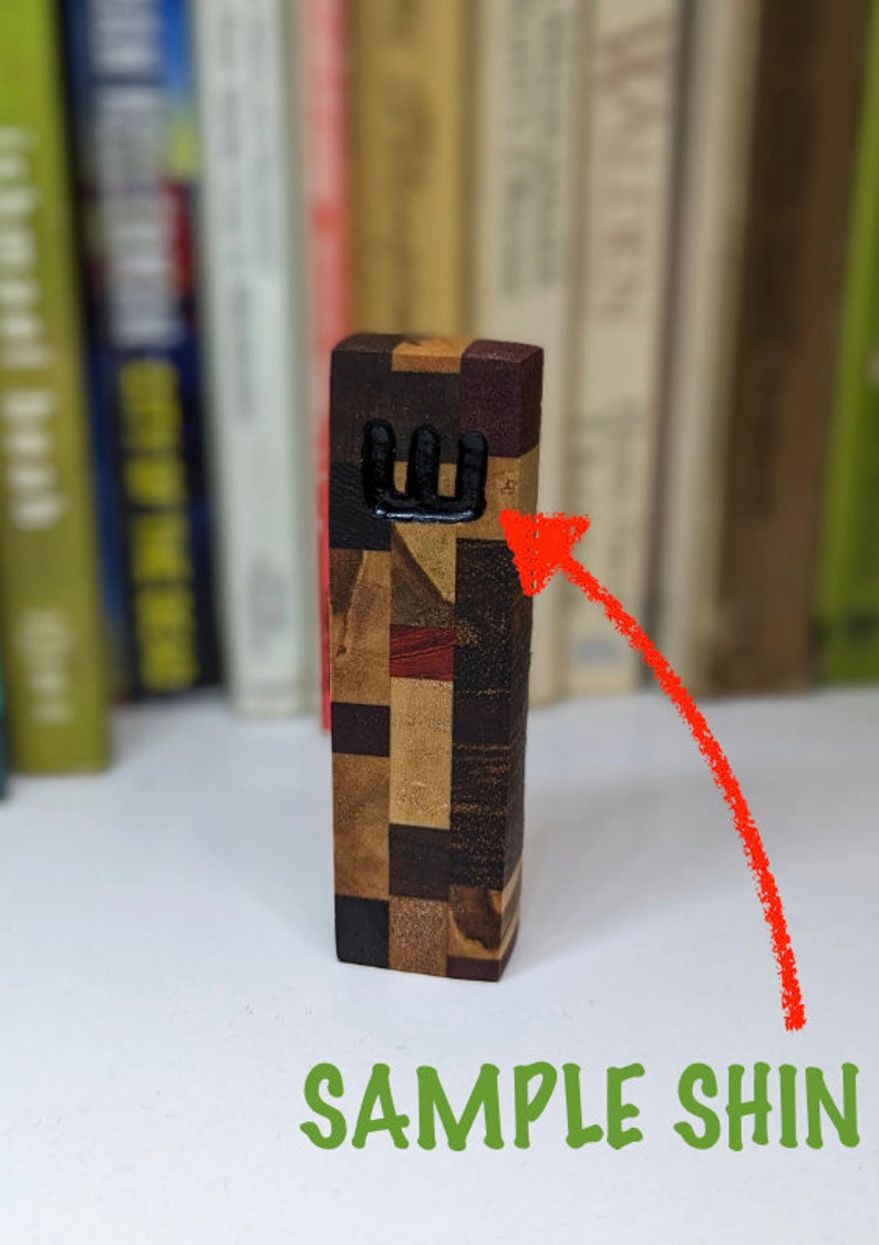 May include: A wooden mezuzah with a black Hebrew letter Shin on the front. The mezuzah is made of different types of wood, creating a patchwork pattern.