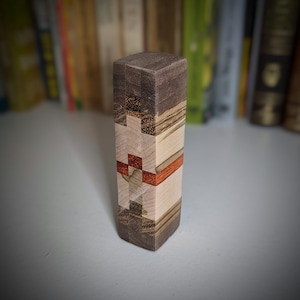 May include: A small wooden block with a multi-coloured wood grain pattern. The block is made of different types of wood, including dark brown, light brown, and red. The block is approximately 7.6 cm tall and 2.5 cm wide.