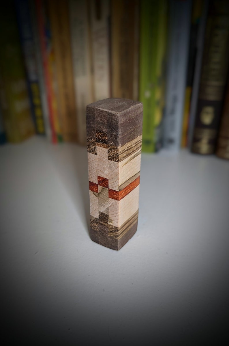 May include: A small wooden block with a multi-coloured wood grain pattern. The block is made of different types of wood, including dark brown, light brown, and red. The block is approximately 7.6 cm tall and 2.5 cm wide.