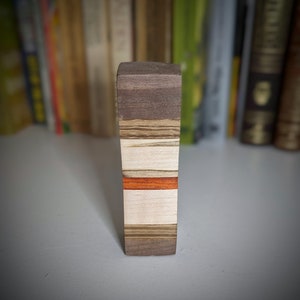 May include: A wooden block with a layered design. The block is made of different types of wood, including walnut, maple, and cherry. The block has a red stripe running through the centre.