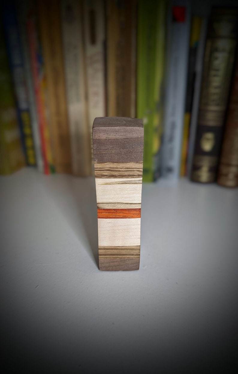 May include: A wooden block with a layered design. The block is made of different types of wood, including walnut, maple, and cherry. The block has a red stripe running through the centre.
