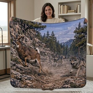 May include: A rectangular throw blanket featuring a high-resolution printed scene of a white-tailed deer with antlers leaping across a rocky, dirt path in a forest, pursued by two grey wolves. The scene includes pine trees and distant mountains.