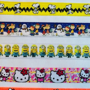 May include: Six different colorful ribbon trims featuring various cartoon characters. The first ribbon is white with a green, blue, and yellow background and features Snoopy characters. The second ribbon is yellow with black zigzags and features Snoopy characters. The third ribbon is blue with white snowflakes and features Snoopy characters. The fourth ribbon is white with a blue background and features Minion characters. The fifth ribbon is white with a pink, yellow, and blue background and features Hello Kitty characters. The sixth ribbon is orange with white flowers and hearts and features Hello Kitty characters.