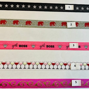 May include: Five different types of ribbon with various patterns. The first ribbon is black with white stars. The second ribbon is silver with red elephants. The third ribbon is pink with a diamond pattern and the text "girl BOSS". The fourth ribbon is white with red wine glasses. The fifth ribbon is pink with a gold swirl pattern.