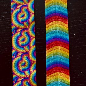 May include: Two colourful fabric strips. One has a repeating pattern of rainbow-coloured swirls. The other displays a rainbow chevron design. The strips are vibrant with hues of red, orange, yellow, green, blue, and purple.