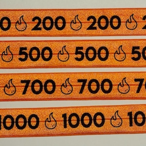 May include: Orange elastic bands with black text and a flame graphic. The bands display numbers: 200, 500, 700, and 1000. The flame graphic is repeated next to each number. The bands are arranged horizontally.