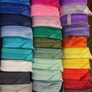 May include: A collection of folded elastic ribbon rolls in a wide spectrum of colors including blue, pink, purple, green, yellow, orange, red, white, gray, black, and brown. Each roll features a textured surface with a distinct diagonal fold line.