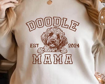 Può includere: Felpa beige con la scritta "DOODLE MAMA EST 2024" in marrone. Un'illustrazione marrone di un cane Doodle è centrata sotto la parola "DOODLE". La felpa è in stile casual.