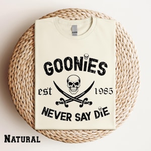 May include: A natural-colored t-shirt featuring a black graphic print with the text GOONIES, est 1985, and NEVER SAY DIE. The design includes a central skull positioned above two crossed swords, with two smaller skulls placed above and below the text.