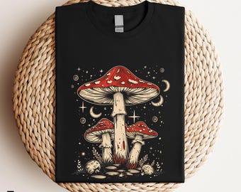 Könnte beinhalten: Ein schwarzes T-Shirt mit einer grafischen Illustration von drei roten und cremefarbenen Pilzen. Das Design enthält himmlische Elemente wie Sterne, Mondsicheln und kleine kreisförmige Muster, die die Pilze auf dunklem Hintergrund umgeben.
