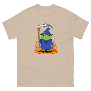 Könnte beinhalten: Beigefarbenes T-Shirt mit einem Cartoon-Frosch als Zauberer, der einen blauen Umhang und Hut trägt, einen Stab hält und in Flammen steht. Der Text "heals pls" steht über dem Frosch.