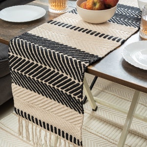 Puede incluir: Camino de mesa tejido con un patrón geométrico negro y crema. El camino de mesa presenta una serie de líneas diagonales y horizontales negras, con un fondo crema. El camino de mesa tiene un borde con flecos y está colocado sobre una mesa de madera.