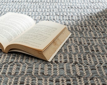 May include: An open book lying on a grey and white patterned rug. The book is open to a page with text.
