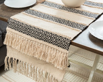 Op de afbeelding: Een geweven tafelkleed in beige en zwart met een geometrisch patroon en franjes. De tafelkleed ligt op een houten tafel met een wit bord en een kom met appels.