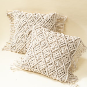 Op de afbeelding: Twee crèmekleurige macramé kussens met franjes. De kussens hebben een ruitpatroon en zijn gemaakt van natuurlijke vezels.