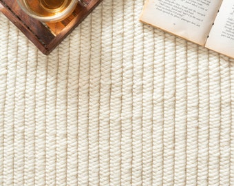 Op de afbeelding: Een wit geweven tapijt met een subtiele textuur. Het tapijt heeft een licht beige kleur en is gemaakt van natuurlijke vezels. Een houten dienblad met een glas thee staat op het tapijt.
