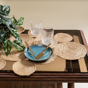 May include: A wooden table with a glass top, set with blue and white ceramic plates, gold cutlery, and woven placemats. Two clear glass tumblers are on the table.