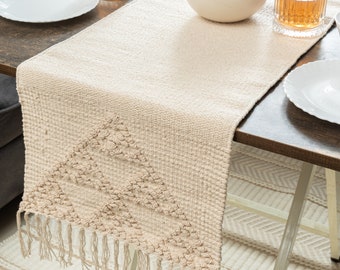 Op de afbeelding: Beige geweven tafelloper met een geometrisch ontwerp en franjes. De loper ligt op een donkerbruine houten tafel, met witte borden, glazen en een kom met rode appels. De tafelloper voegt textuur en stijl toe aan de tafel.