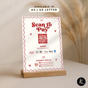 May include: A white sign with red text and a QR code, displayed on a wooden stand. The sign reads "Scan to Pay" and includes payment options. The sign also says "Connect with us" with social media handles.