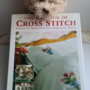May include: A book titled "The Big Book of Cross Stitch" with a white background and red text. The cover features a photo of a white bed with a light blue sheet and a cross-stitch design of pink and blue flowers. The book is by Jan Eaton and Dorothea Hall.