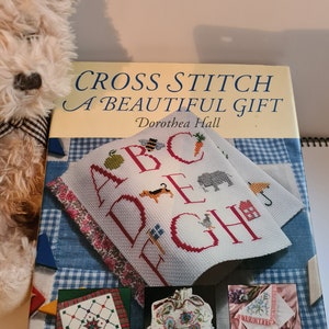 May include: A book titled "Cross Stitch A Beautiful Gift" by Dorothea Hall. The cover features a cross stitch design of the alphabet with various animals and objects. The book also shows three smaller cross stitch designs: a cushion, a bag, and a handkerchief.