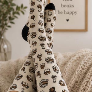 May include: Cream-colored crew socks featuring a repeating pattern of brown and black illustrations of stacked books and coffee-related items like portafilters. The socks have solid black toes and heels, providing a contrast to the light base color.