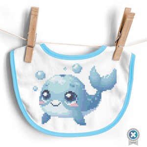 May include: A white baby bib with a blue border and a blue and white embroidered design of a cartoon whale with big eyes and bubbles.