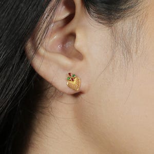 May include: A gold-colored heart-shaped stud earring with a detailed, textured design. The earring features red and green gemstone accents at the top. The earring is worn on the ear of a person.