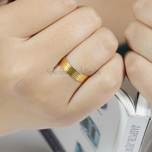 Puede incluir: Un anillo de banda de oro amarillo pulido que presenta una serie de cinco ranuras horizontales y paralelas grabadas en la superficie. El anillo se lleva en un dedo, mostrando un acabado metálico liso y reflectante con un ancho uniforme y constante en toda la pieza.