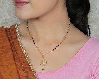 May include: A gold-colored necklace with a pendant and black beads. The pendant has an ornate design with small black beads hanging from it. The necklace is worn on a person wearing a pink top and an orange shawl.