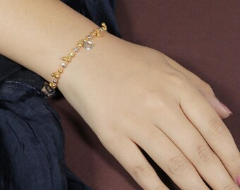 May include: A gold-toned bracelet with a delicate design featuring small, textured beads and a dangling clear charm. The bracelet is worn on a wrist with pink nail polish, set against a dark background.