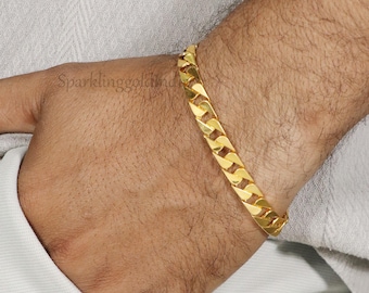 May include: A gold curb chain bracelet featuring interlocking flat, polished links. The bracelet has a bright, reflective yellow gold finish and a classic, uniform design that wraps around the wrist. The metal surface is smooth and highly reflective.