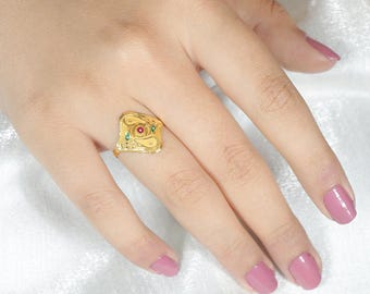 May include: A gold-colored ring with a square face, featuring a central red stone and smaller green stones. The ring has an ornate design with swirling patterns. The ring is on a hand with pink nail polish.