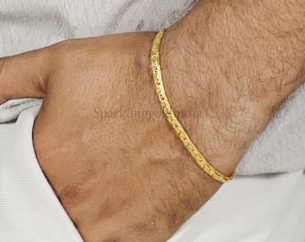May include: A thin, polished gold bracelet with a textured, repeating link pattern worn around the wrist. The metal surface reflects light, highlighting the intricate, interconnected design of the chain links that form a continuous, flexible band.