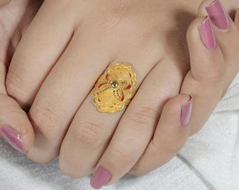 May include: A gold ring featuring an oval-shaped face with intricate filigree metalwork, scalloped edges, and a small, centered green gemstone. The design includes delicate cutouts and textured patterns across the surface of the metal.