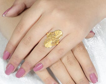 May include: A gold-colored ring with an intricate floral design. The ring features a central flower with detailed petals and surrounding openwork patterns. The ring is worn on a finger, with the hands and nails painted with a mauve nail polish.