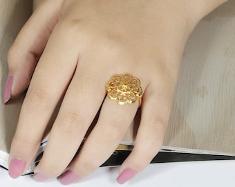 May include: A gold-colored ring with a floral design. The ring features an intricate, openwork pattern with a central, raised element. The ring is worn on a finger, with the hand resting on a surface.