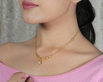 May include: A gold-colored necklace with a delicate design. The necklace features a central pendant and ornate detailing along the chain. The necklace is worn on a person wearing a pink top.