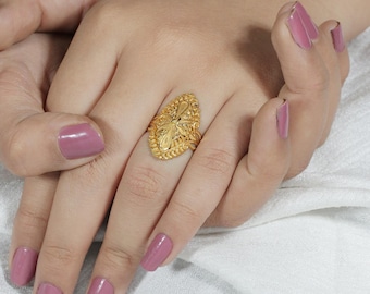 May include: A gold-colored ring with an oval shape and intricate floral and leaf detailing. The ring is worn on a finger, showcasing its ornate design and polished finish. The ring's design includes a central floral element and a textured band.