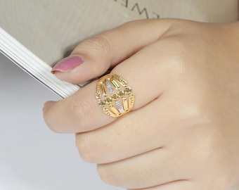 May include: A gold-colored ring with an openwork design featuring vertical bars and small, round accents. The ring has a central section with small, clear stones. The ring is on a person's finger.
