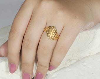 May include: A gold ring with a lattice design. The ring features a large, oval-shaped face with a grid pattern. The ring is worn on a finger, and the hand is resting on a white surface. The ring is made of a shiny, yellow metal.