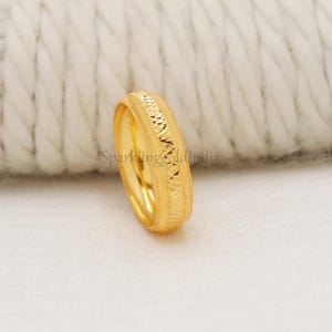 Könnte beinhalten: Ein goldener Ring mit strukturiertem Design. Der Ring hat ein Band mit einem sich wiederholenden geometrischen Muster und einer polierten Oberfläche. Der Ring ist vor einem weißen Hintergrund platziert.