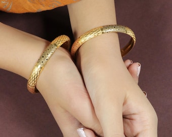 Könnte beinhalten: Zwei goldene Armreifen mit aufwendigen geometrischen Mustern werden gezeigt. Die Armreifen haben eine strukturierte Oberfläche und werden an Handgelenken getragen. Der Schmuck hat eine warme Goldfarbe, und das Design enthält schwarze Akzente. Der Hintergrund ist einfarbig braun.
