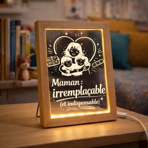 Op de afbeelding: Houten LED-verlichte lijst met de woorden "Maman: irremplaçable (et indispensable)" en een afbeelding van een moeder die twee kinderen omhelst. Het licht heeft een warme gloed en een wit netsnoer.
