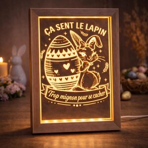 Peut inclure: Cadre lumineux sur le thème de Pâques, avec un design de lapin et d'œuf. Le texte "Ça sent le lapin" et "Trop mignon pour se cacher" est visible. Le cadre en bois rectangulaire diffuse une lumière chaleureuse.