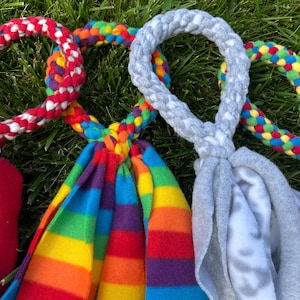 May include: Close-up of several dog toys on green grass. The toys feature colorful braided rope handles and knotted fleece tails in various colors, including red, rainbow, and gray.