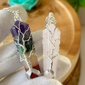 May include: Two wire-wrapped crystal pendants, one with a multi-coloured stone and the other with a pink stone. The pendants are on a silver chain and are displayed on a wooden surface. @shifatagate